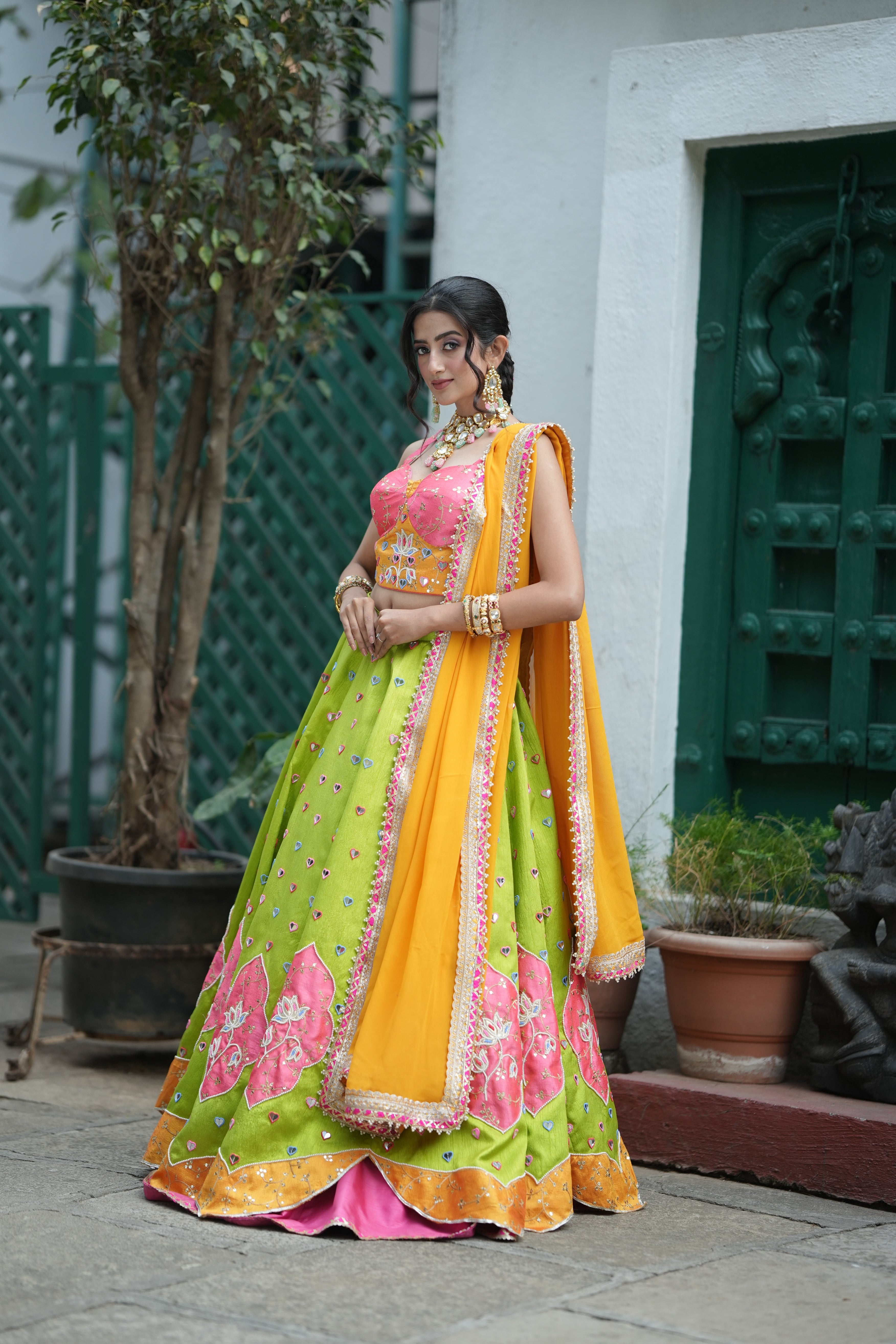 Woman wearing a vibrant lime green Multi-Colour Mirror Work lehenga with floral embroidery, pink blouse, and orange dupatta, posing outdoors.